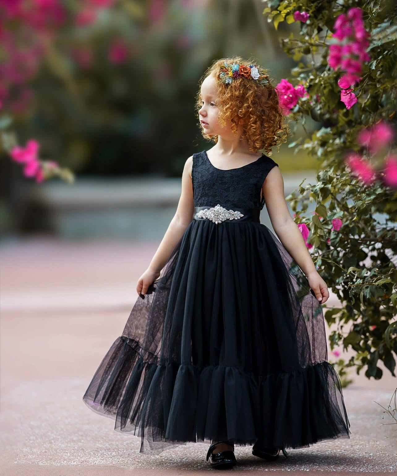 Little girl wearing a sleeveless black lace and tulle mermaid-style dress with a jeweled sash, standing near pink flowers and wearing a colorful floral headband.