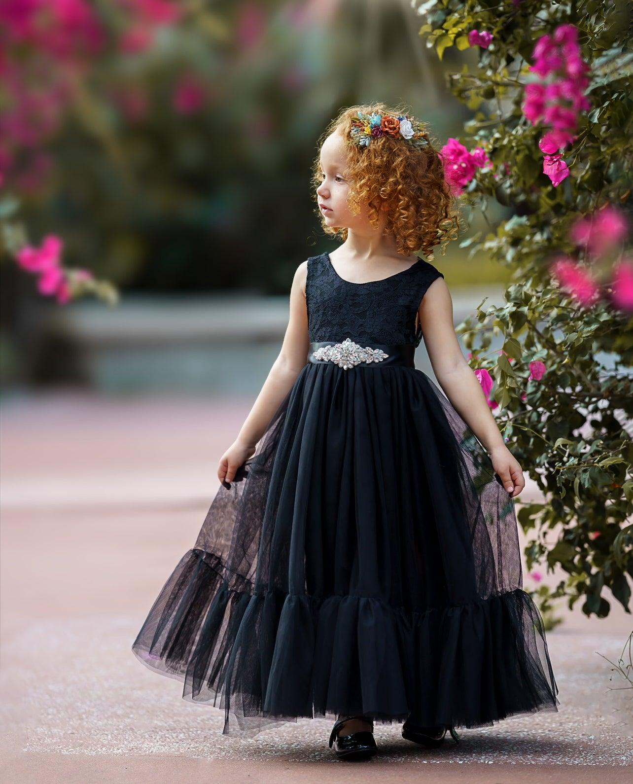 Little girl wearing a sleeveless black lace and tulle mermaid-style dress with a jeweled sash, standing near pink flowers and wearing a colorful floral headband.
