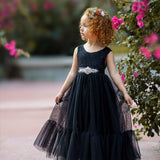Little girl wearing a sleeveless black lace and tulle mermaid-style dress with a jeweled sash, standing near pink flowers and wearing a colorful floral headband.