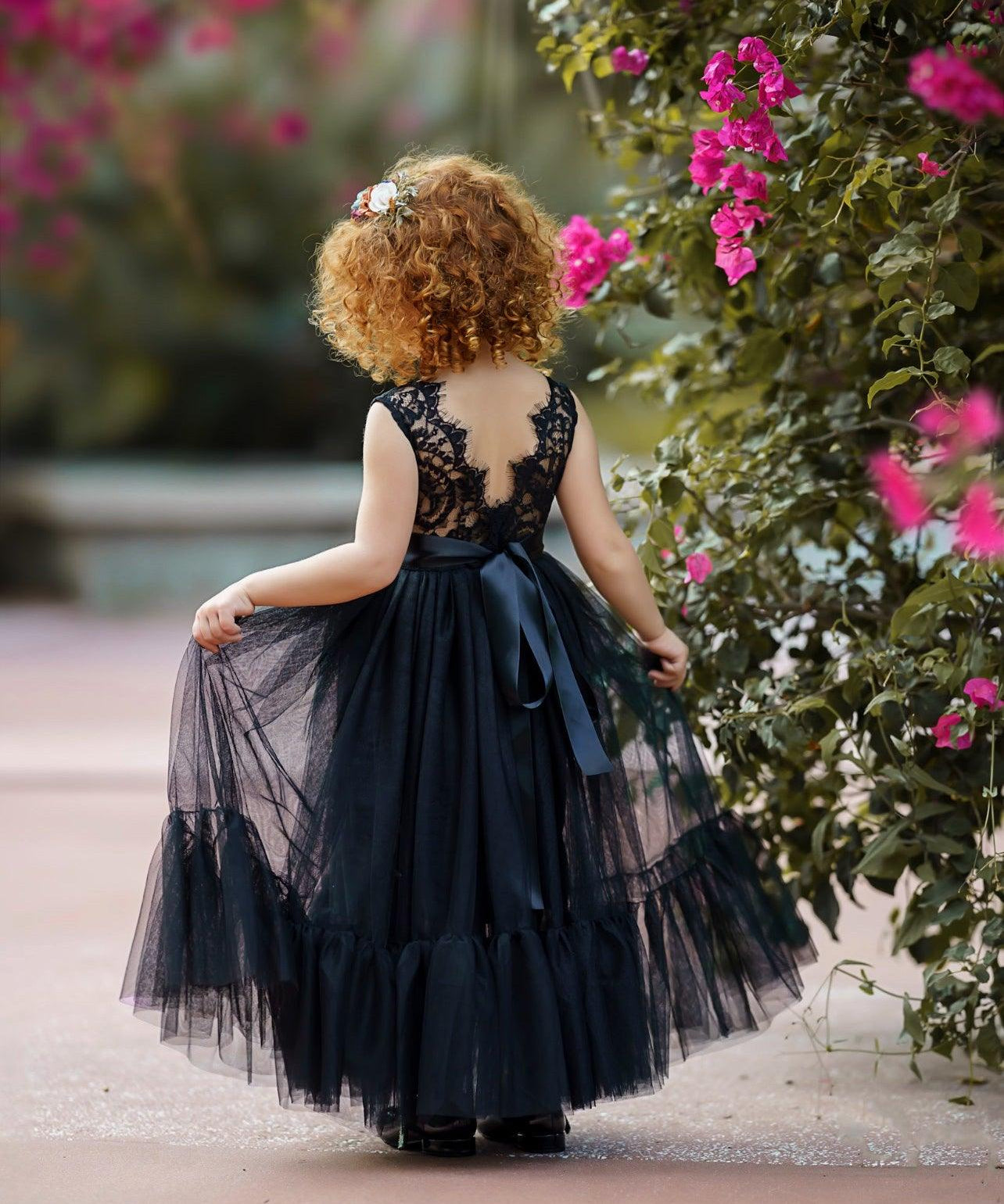 Young girl wearing a sleeveless black lace and tulle mermaid-style flower girl dress with a satin sash, standing outdoors among blooming pink flowers with a floral headpiece.