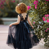 Young girl wearing a sleeveless black lace and tulle mermaid-style flower girl dress with a satin sash, standing outdoors among blooming pink flowers with a floral headpiece.