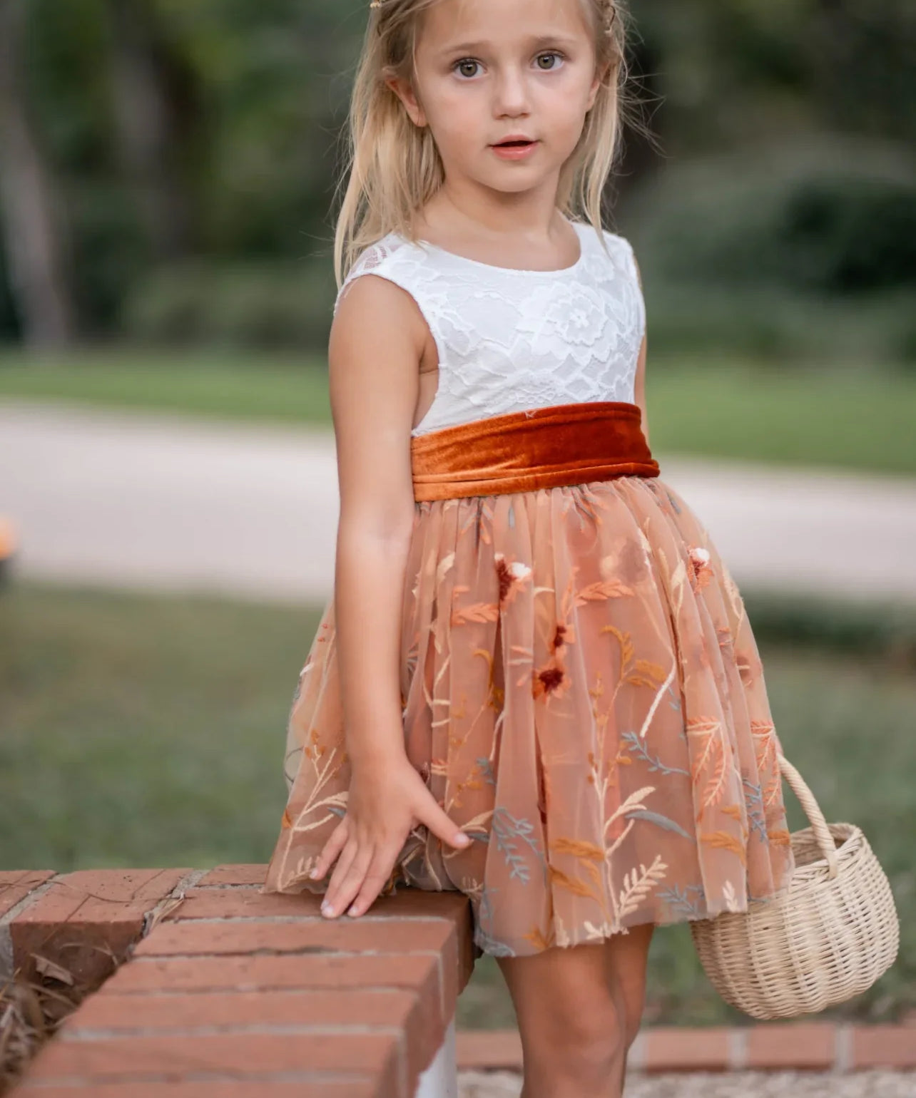 Front view of girl wearing burnt orange floral embroidered flower girl dress with white lace bodice and velvet sash