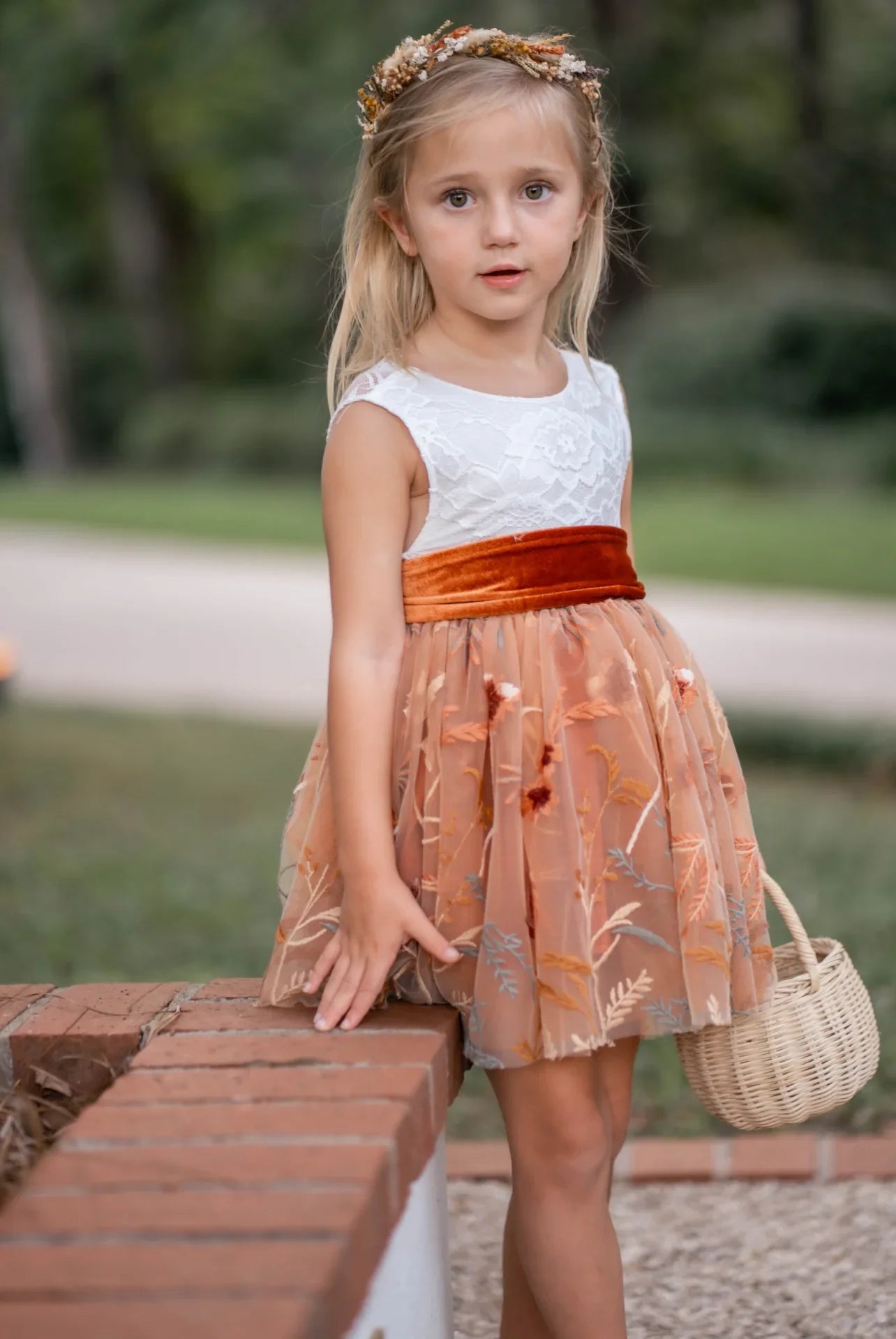 Front view of girl wearing burnt orange floral embroidered flower girl dress with white lace bodice and velvet sash